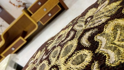 An intricate fabric with golden and brown embroidery in the foreground, with a background of wooden furniture featuring geometric designs.