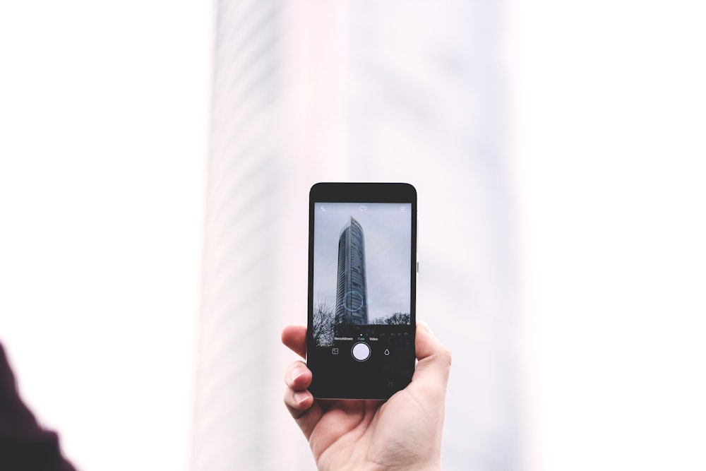 Fotografia a fuoco superficiale di persona che scatta foto della torre di vetro allo smartphone