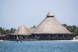 A large thatched-roof building, resembling a traditional hut, stands near a sandy beach with palm trees in the background. In the foreground, a small sailboat with a colorful sail is being operated by a person on the water.