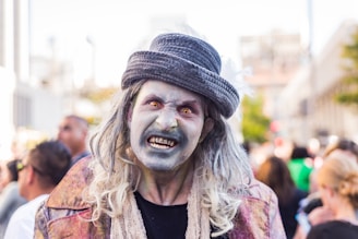 A person dressed as a zombie in a public setting, featuring pale makeup and exaggerated features to create a frightening appearance. They wear a hat and have long hair with clothing resembling tattered attire, indicating a costume for a horror-themed event.