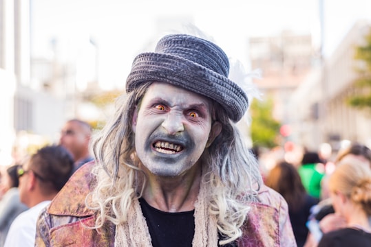 A person dressed as a zombie in a public setting, featuring pale makeup and exaggerated features to create a frightening appearance. They wear a hat and have long hair with clothing resembling tattered attire, indicating a costume for a horror-themed event.