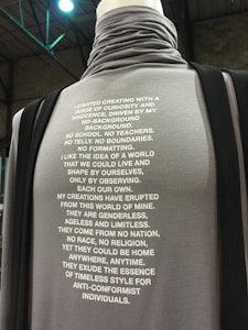 A mannequin is displayed wearing a high-collared grey garment featuring a lengthy paragraph written in bold white text. This text elaborates on a philosophy of universal, boundary-breaking creativity. The neck and upper chest are tucked, emphasizing the text's presentation.