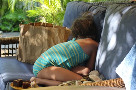 A child wearing a striped swimsuit is curled up on a cushioned outdoor sofa, facing away. Next to the child, on the sofa, is a brown paper bag and some food items. The surroundings include green foliage, and sunlight casts dappled shadows on the scene.