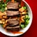 A bowl filled with grilled meat slices, cashew nuts, edamame beans, leafy greens, carrots, and other vegetables. The presentation is vibrant and colorful, with fresh ingredients arranged aesthetically.