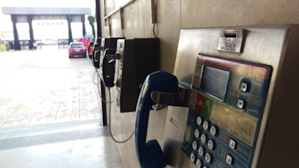 A series of vintage payphones are mounted on a wall in a semi-outdoor setting. One of the payphones in the foreground has a blue handset and visible coin slot. A red car is parked in the background near a gated entrance.