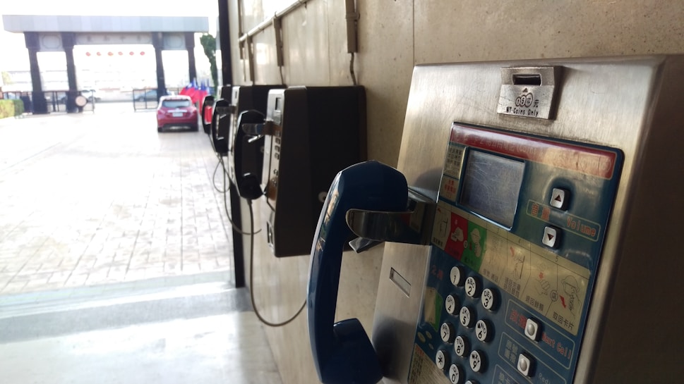 A series of vintage payphones are mounted on a wall in a semi-outdoor setting. One of the payphones in the foreground has a blue handset and visible coin slot. A red car is parked in the background near a gated entrance.