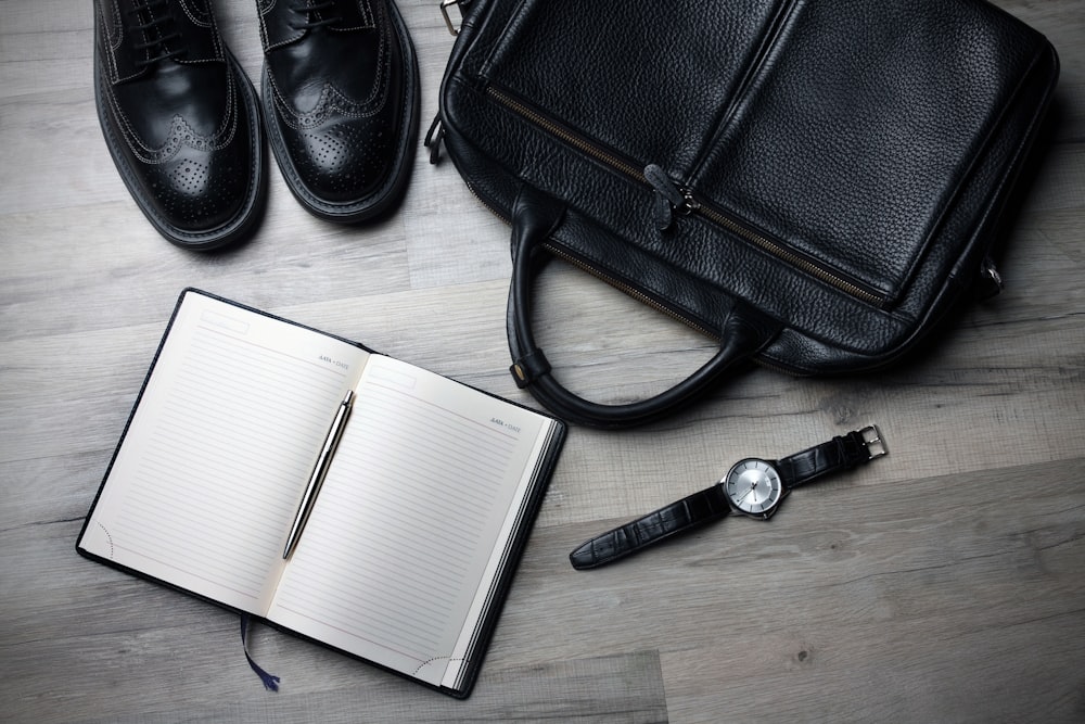 An overhead shot of a leather bag, a pair of leather shoes and a watch next to an open notebook