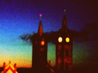 Two dark, pointed towers set against a richly colored gradient sky transitioning from blue to orange, suggesting twilight or dawn. The towers have glowing round lights resembling eyes, contributing to a slightly eerie atmosphere.