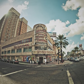 A corner street view featuring an old Walgreens store with large vintage signage advertising drugs, cosmetics, and photo services. The sunlight creates a warm atmosphere, casting shadows on the building. Palm trees line the street alongside several parked cars, and pedestrians are visible on the sidewalk. The scene is set against a backdrop of taller buildings and a partly cloudy sky.