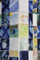 A collection of ceramic tiles featuring abstract and colorful designs. The patterns include splashes of blue, green, yellow, and various other colors, with geometric and freeform shapes. The tiles are arranged in a grid pattern on a wall, creating a mosaic-like appearance.