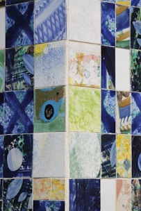 A collection of ceramic tiles featuring abstract and colorful designs. The patterns include splashes of blue, green, yellow, and various other colors, with geometric and freeform shapes. The tiles are arranged in a grid pattern on a wall, creating a mosaic-like appearance.
