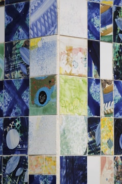 A collection of ceramic tiles featuring abstract and colorful designs. The patterns include splashes of blue, green, yellow, and various other colors, with geometric and freeform shapes. The tiles are arranged in a grid pattern on a wall, creating a mosaic-like appearance.