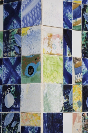 A collection of ceramic tiles featuring abstract and colorful designs. The patterns include splashes of blue, green, yellow, and various other colors, with geometric and freeform shapes. The tiles are arranged in a grid pattern on a wall, creating a mosaic-like appearance.