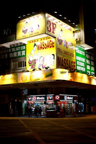 A brightly lit building at night features prominent yellow signs advertising a Thai massage parlor on the third floor. The signage includes illustrations of people receiving massages and contact information. Below, a convenience store named Circle K is illuminated, showing people entering and leaving.