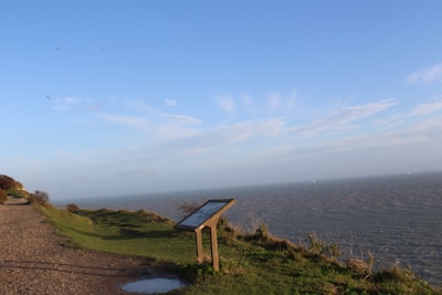 A coastal landscape with a grassy path running alongside the ocean. An informational sign on a stand is positioned near the edge of the cliff. The sky is clear with a few scattered clouds and several birds flying in the distance.