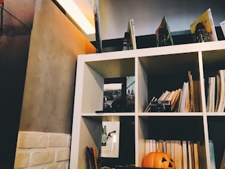 A cozy interior space featuring a white shelf filled with books, magazines, and a vintage camera. A carved pumpkin decoration sits on the bottom row, adding a festive touch. The wall behind the shelf is a combination of beige textured paint and white brick, with soft lighting creating a comfortable ambiance.