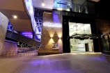 An architectural scene showcasing the entrance to a modern public building with prominent brick walls and large glass doors under nighttime lighting. The building is illuminated with purple and white lighting, reflecting an urban, contemporary design. The entrance is situated under a canopy structure and there are metal railings leading to the building.