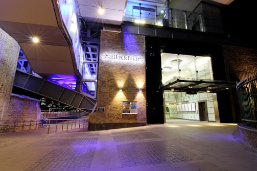 An architectural scene showcasing the entrance to a modern public building with prominent brick walls and large glass doors under nighttime lighting. The building is illuminated with purple and white lighting, reflecting an urban, contemporary design. The entrance is situated under a canopy structure and there are metal railings leading to the building.