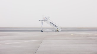 A mobile stairway is positioned on the empty tarmac of an airport, with a foggy and minimalistic backdrop. The stairway features branding and wheels at its base, sitting on an expansive, barren runway.