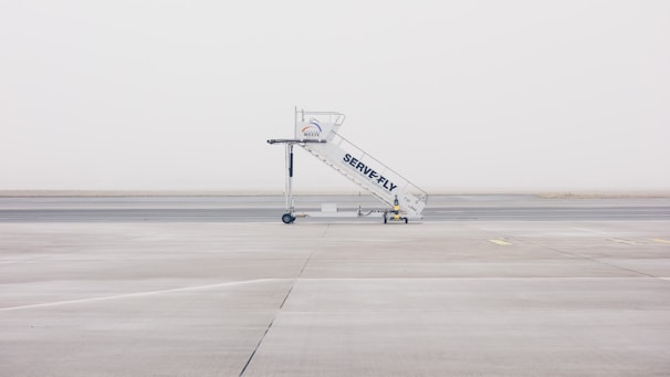 A mobile stairway is positioned on the empty tarmac of an airport, with a foggy and minimalistic backdrop. The stairway features branding and wheels at its base, sitting on an expansive, barren runway.