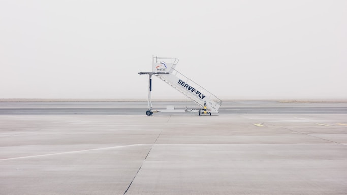 A mobile stairway is positioned on the empty tarmac of an airport, with a foggy and minimalistic backdrop. The stairway features branding and wheels at its base, sitting on an expansive, barren runway.