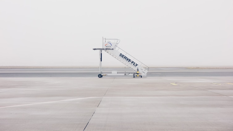 A mobile stairway is positioned on the empty tarmac of an airport, with a foggy and minimalistic backdrop. The stairway features branding and wheels at its base, sitting on an expansive, barren runway.