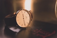 A classic wristwatch with a minimalist design featuring a white dial, thin black indices, and sleek black hands. It has a brown leather strap and is placed on a dark glossy surface with soft, warm lighting creating a gentle reflection. A red insignia or mark is visible on the surface near the watch.