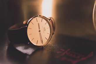 A classic wristwatch with a minimalist design featuring a white dial, thin black indices, and sleek black hands. It has a brown leather strap and is placed on a dark glossy surface with soft, warm lighting creating a gentle reflection. A red insignia or mark is visible on the surface near the watch.