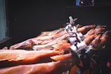 A display of cured ham legs secured in metal holders, showcasing the texture and color of the meat. The meats vary in shade, highlighting marbling and an array of warm tones.