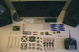 A neatly organized workstation displays various drone parts and tools laid out on a desk. Components include blue propellers, electronic circuit boards, batteries, connectors, and a remote controller. The setup is complemented by a large monitor, keyboard, and mouse. Two speakers are placed on either side.