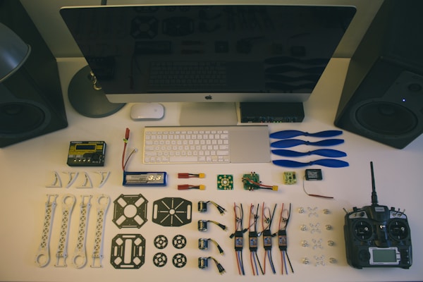 A neatly organized workstation displays various drone parts and tools laid out on a desk. Components include blue propellers, electronic circuit boards, batteries, connectors, and a remote controller. The setup is complemented by a large monitor, keyboard, and mouse. Two speakers are placed on either side.