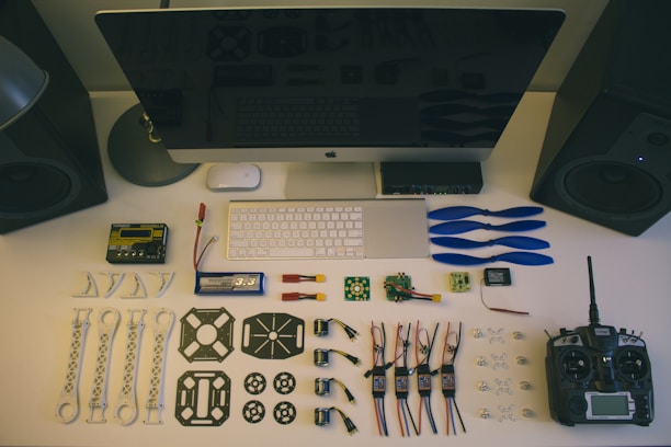 A neatly organized workstation displays various drone parts and tools laid out on a desk. Components include blue propellers, electronic circuit boards, batteries, connectors, and a remote controller. The setup is complemented by a large monitor, keyboard, and mouse. Two speakers are placed on either side.