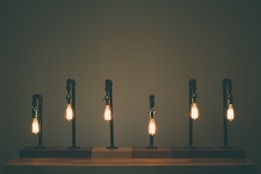 A row of six vintage-style light bulbs attached to dark metal fixtures casts a warm glow in an otherwise dimly lit space. The bulbs are evenly spaced on a wooden surface, providing an industrial yet cozy aesthetic.