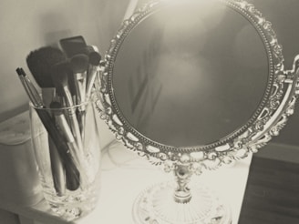 A vintage-style ornate table mirror with intricate designs rests on a surface. Next to it is a tall, clear glass holding various makeup brushes with dark bristles.