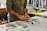 A person wearing a long-sleeved shirt is working at a cluttered desk with various tools and materials scattered around. Their hands are focused on crafting or assembling something using red material. In the background, there are shelves and containers with additional items.