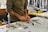 A person wearing a long-sleeved shirt is working at a cluttered desk with various tools and materials scattered around. Their hands are focused on crafting or assembling something using red material. In the background, there are shelves and containers with additional items.