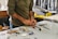 A person wearing a long-sleeved shirt is working at a cluttered desk with various tools and materials scattered around. Their hands are focused on crafting or assembling something using red material. In the background, there are shelves and containers with additional items.