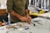 A person wearing a long-sleeved shirt is working at a cluttered desk with various tools and materials scattered around. Their hands are focused on crafting or assembling something using red material. In the background, there are shelves and containers with additional items.