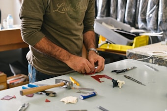 A person wearing a long-sleeved shirt is working at a cluttered desk with various tools and materials scattered around. Their hands are focused on crafting or assembling something using red material. In the background, there are shelves and containers with additional items.