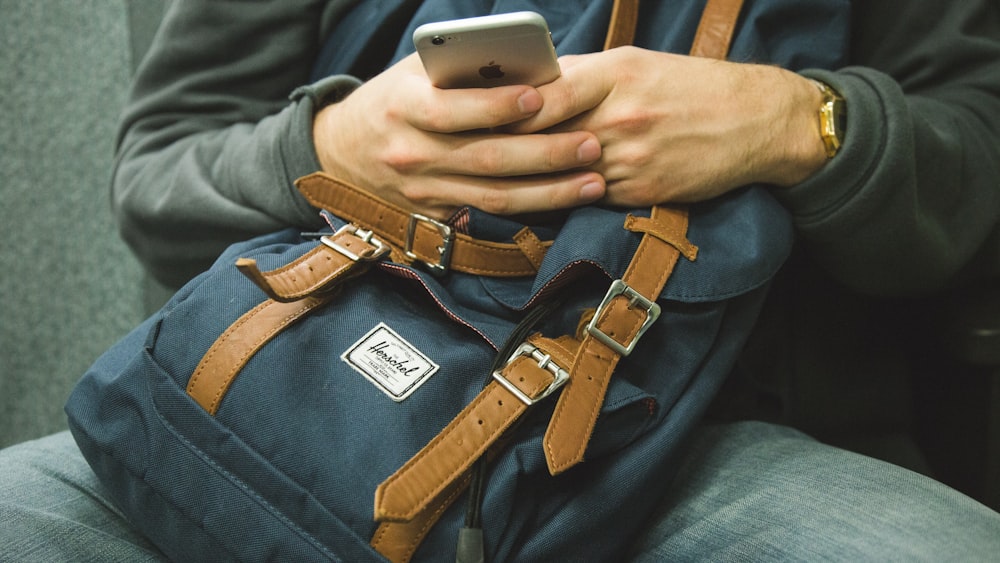 A man with a blue backpack on his lap using an iPhone