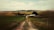 A rural landscape with a dirt road leading through green and brown fields towards a small group of buildings and trees. Rolling hills are visible in the background under a partly cloudy sky.