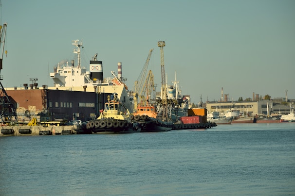 A bustling port area featuring several docked ships and tugboats. A large cargo vessel is prominent in the background, with industrial buildings and cranes creating a busy atmosphere. Shipping containers are stacked along the dock, and the water is calm under a clear sky.