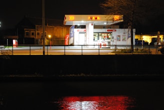 A gas station is illuminated at night, showcasing bright lights against the dark surroundings. The station has a small shop attached, with visible signs and posters. The forecourt is empty, with no visible cars or people. The reflection of the gas station's lights can be seen on the surface of a body of water in the foreground.