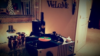 A cozy room features a vintage record player spinning a vinyl next to a bottle of wine and two copper mugs. A candle is placed nearby, and a cocktail shaker sits on the counter. A large mirror reflects a decorated Christmas tree. The word 'Welcome' is displayed on the wall.
