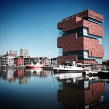 A modern, uniquely-stacked red brick building with large glass windows is situated near a body of water. Several boats are docked along the water's edge, and a smaller boat with a prominent 'West-Hinder' caption on the side can be seen. The waterfront is lined with various buildings, ranging from modern high-rises to older, more traditional structures. The water reflects the buildings and the clear, blue sky above.