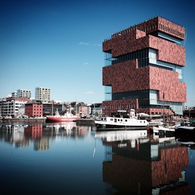 A modern, uniquely-stacked red brick building with large glass windows is situated near a body of water. Several boats are docked along the water's edge, and a smaller boat with a prominent 'West-Hinder' caption on the side can be seen. The waterfront is lined with various buildings, ranging from modern high-rises to older, more traditional structures. The water reflects the buildings and the clear, blue sky above.