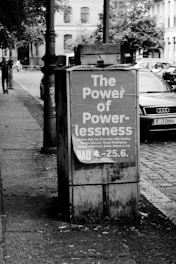 A black and white photograph of a utility box on a street corner displaying a poster with the text 'The Power of Powerlessness' along with some names and a date range. The background shows parked cars, trees, and older buildings.
