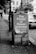 A black and white photograph of a utility box on a street corner displaying a poster with the text 'The Power of Powerlessness' along with some names and a date range. The background shows parked cars, trees, and older buildings.