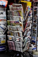 A rack filled with a variety of newspapers and magazines, featuring multiple languages and diverse topics. The publications are neatly arranged in several tiers, showcasing different headlines and imagery. The setting appears to be a newsstand, possibly located in a public area with other items partially visible in the background.