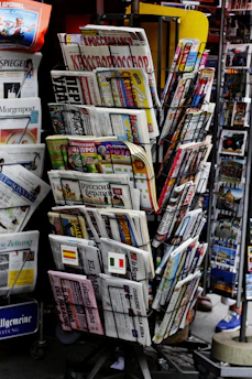 A rack filled with a variety of newspapers and magazines, featuring multiple languages and diverse topics. The publications are neatly arranged in several tiers, showcasing different headlines and imagery. The setting appears to be a newsstand, possibly located in a public area with other items partially visible in the background.